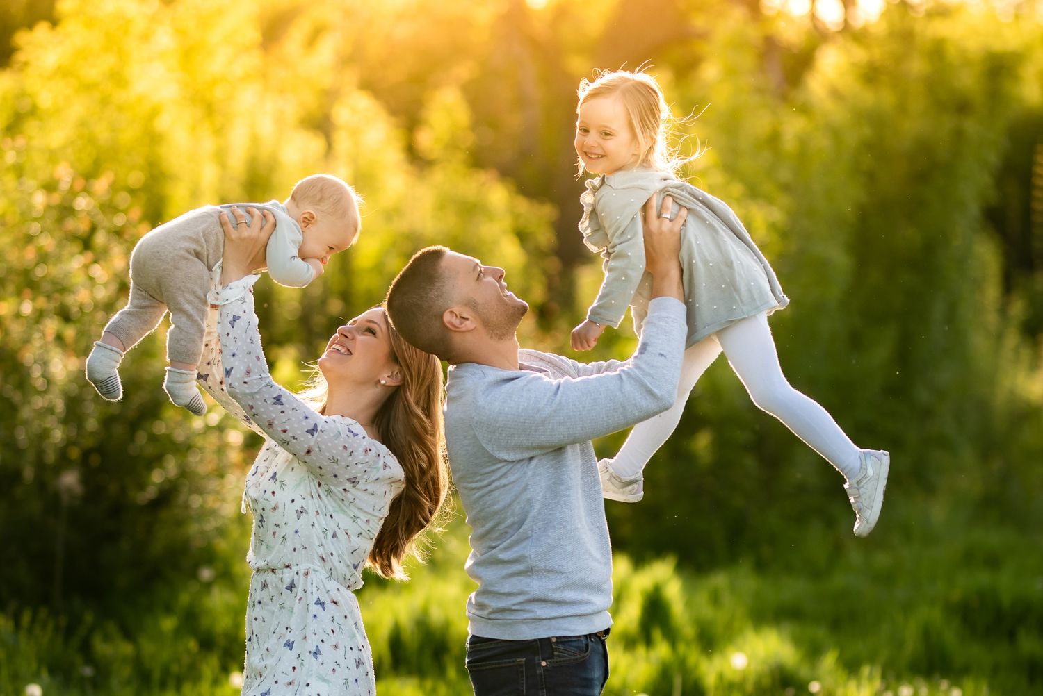 Happy family with parents lifting two young children outdoors in sunny green park setting.