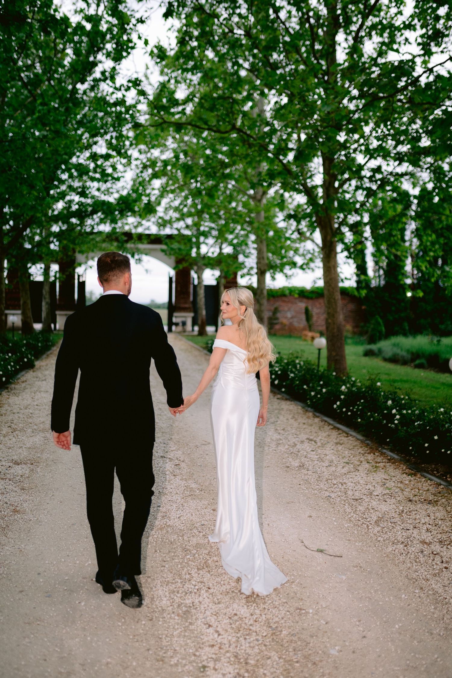 Bride and groom walking hand in hand down a tree-lined gravel path, bride in white off-shoulder gown glancing back.