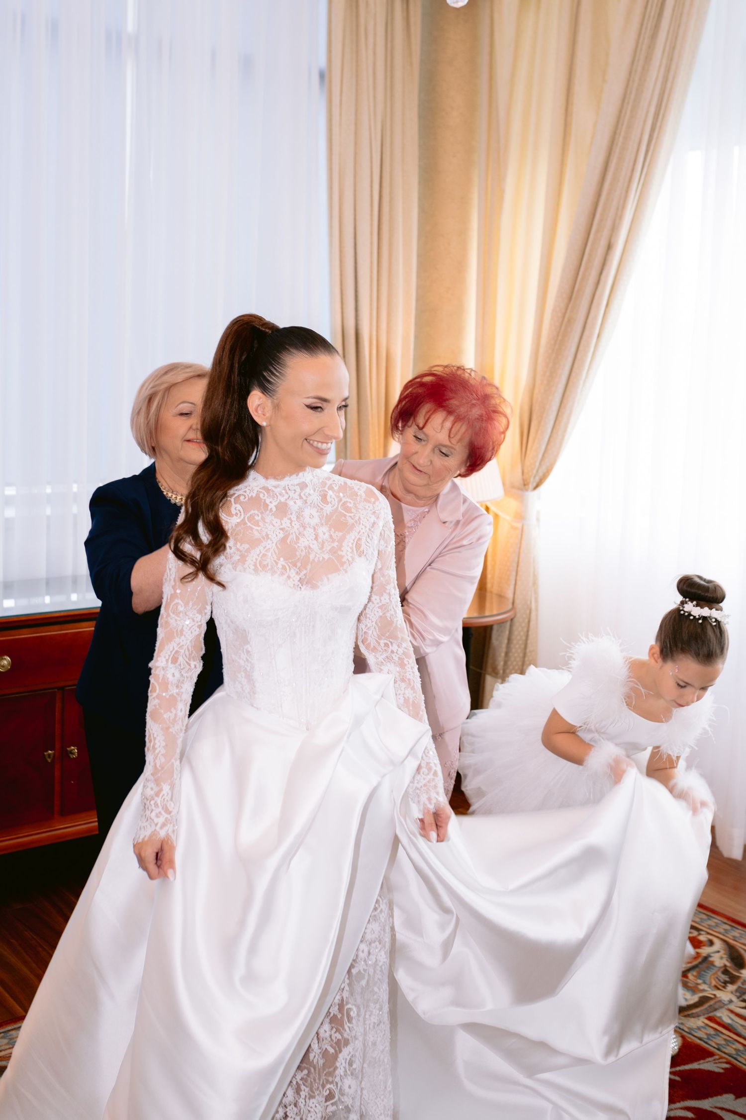 A group of women in white dresses help arrange a wedding gown's long train in an elegant indoor setting.