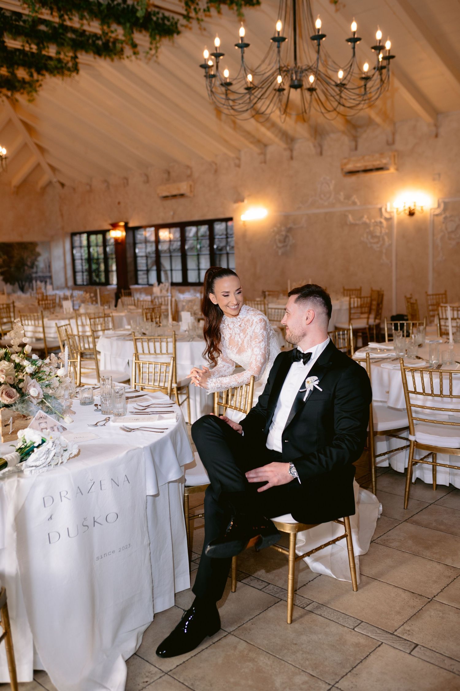 Elegant wedding reception venue with chandelier, white decor, gold chairs and romantic lighting in stone-walled room.