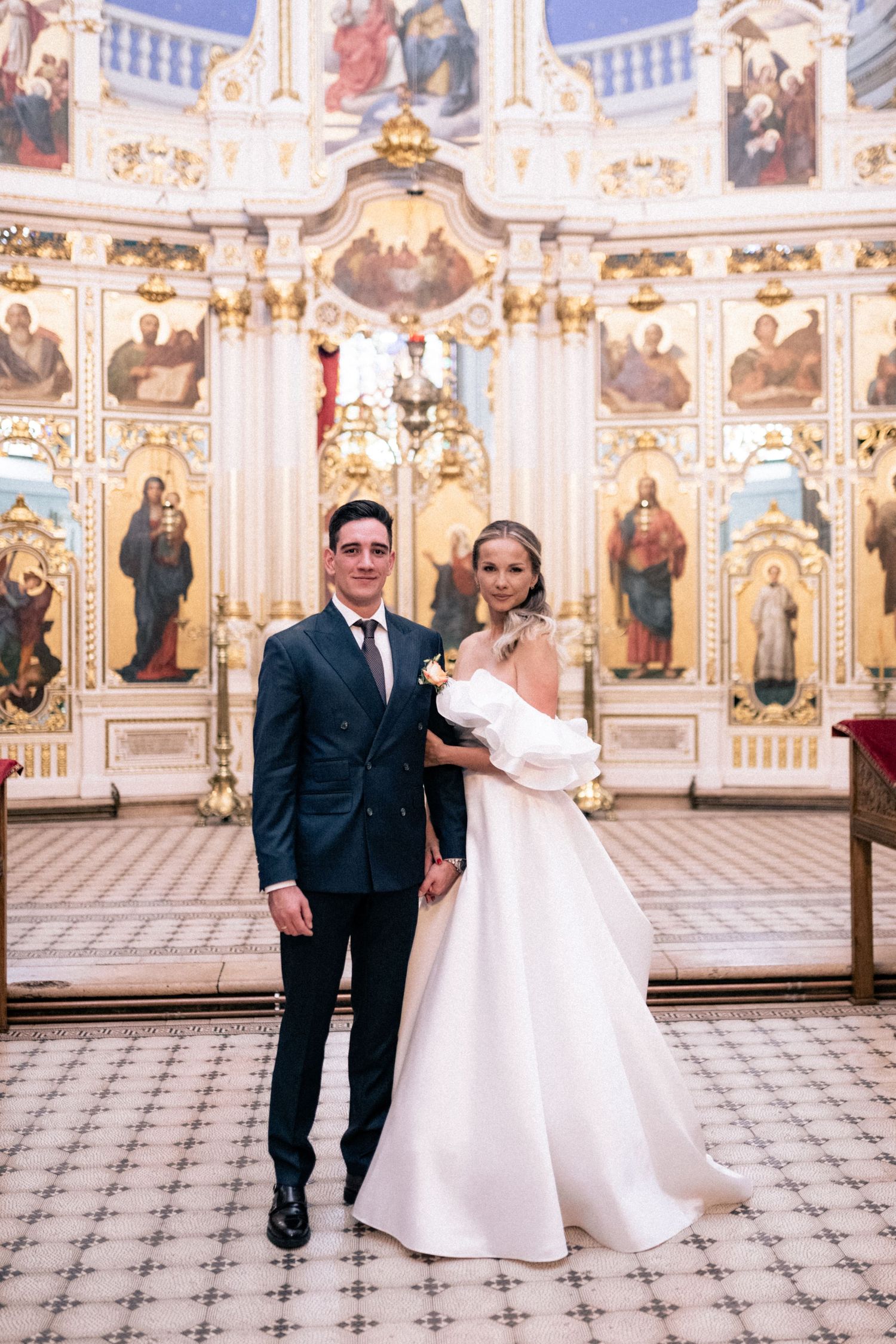 A wedding couple poses in an ornate church interior with gilded baroque decorations and religious paintings on the walls.
