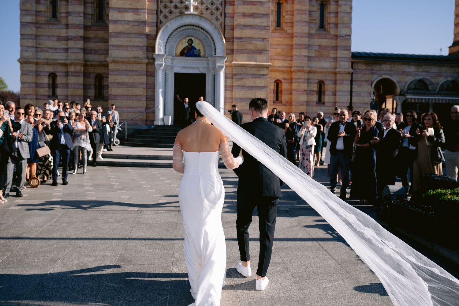 A long flowing veil trails behind a bride in a white dress as wedding guests gather outside a brick church entrance.