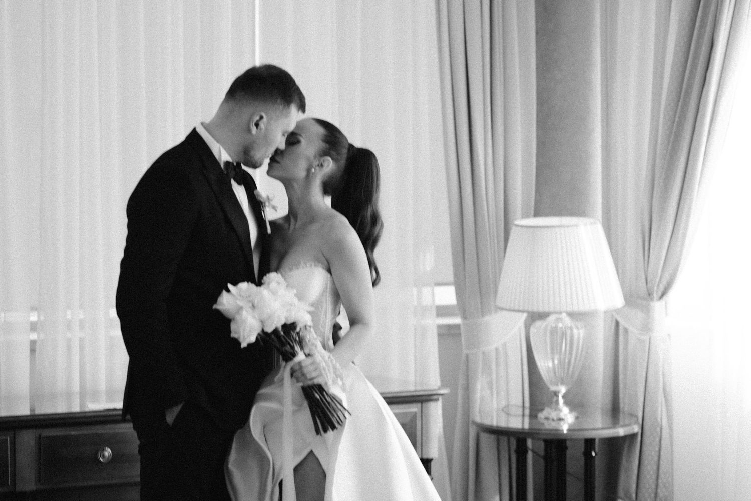 Romantični trenutak mladenaca sa vjenčanja zabilježen u elegantnoj sobi sa bijelim zavjesama.