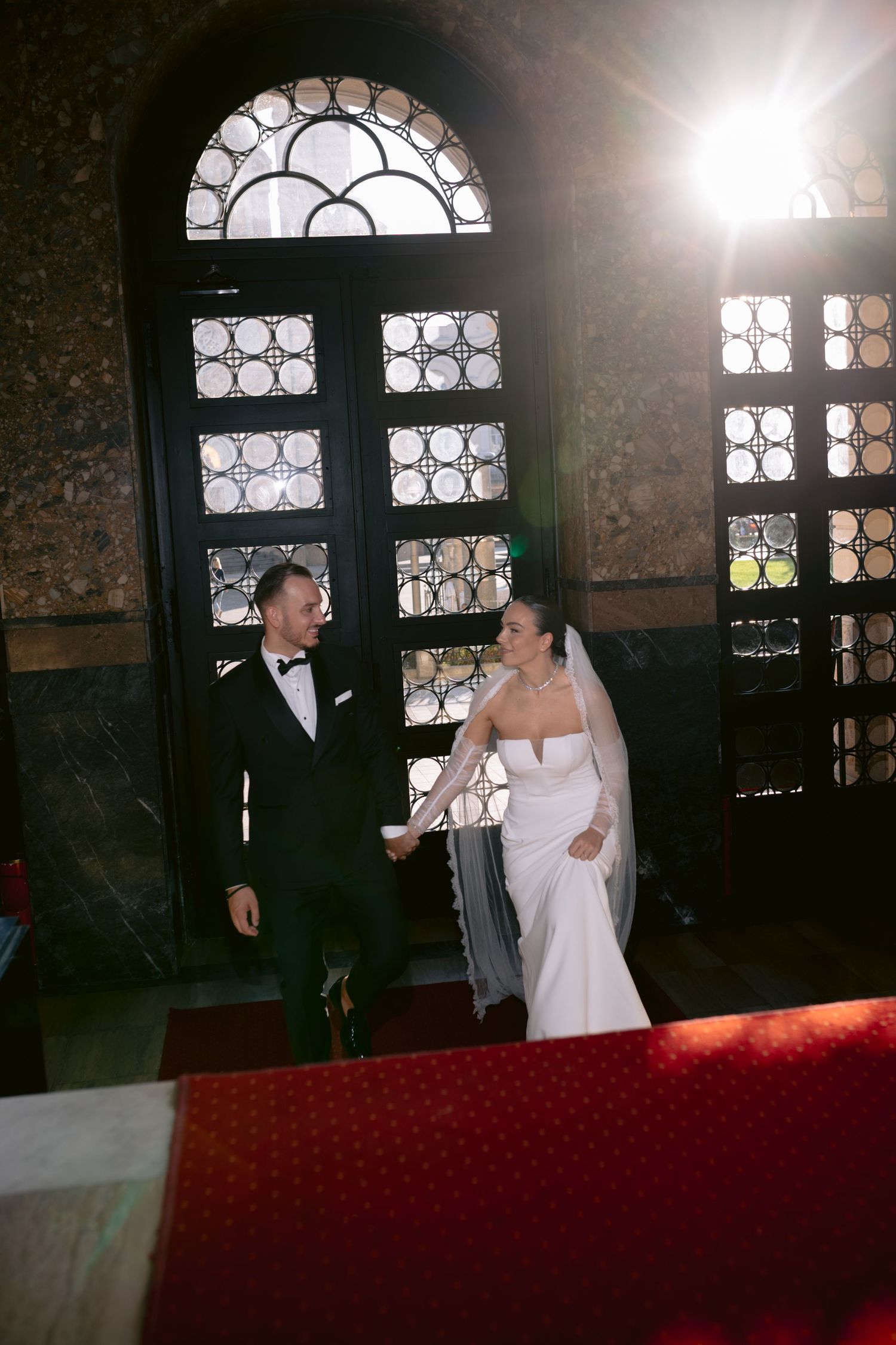 A couple in wedding attire stands in a dimly lit church with ornate stained glass windows and red carpet.