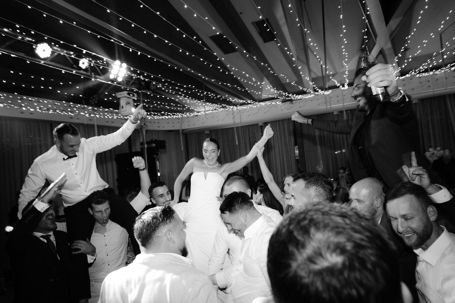 People dancing and celebrating at a wedding reception under decorative string lights and a draped ceiling.