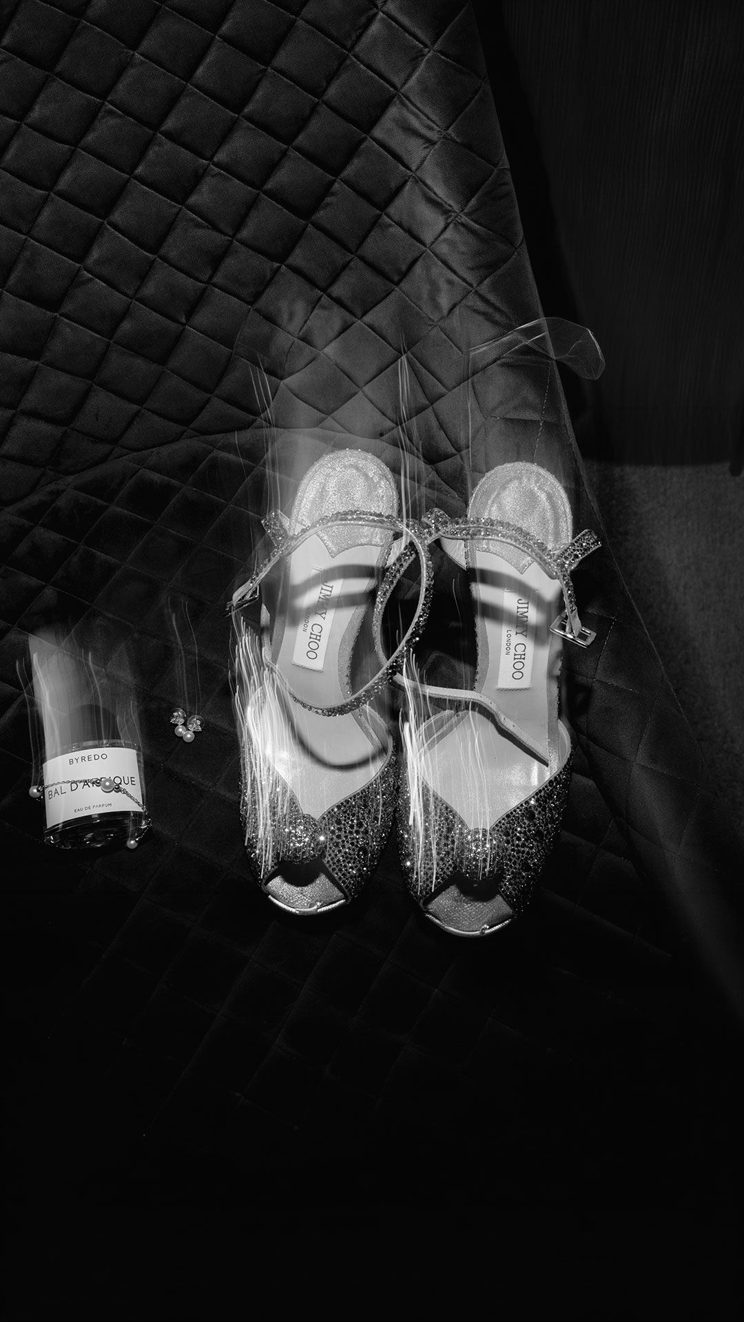 A pair of elegant silver Mary Jane shoes with sparkly details photographed in black and white against a quilted background.