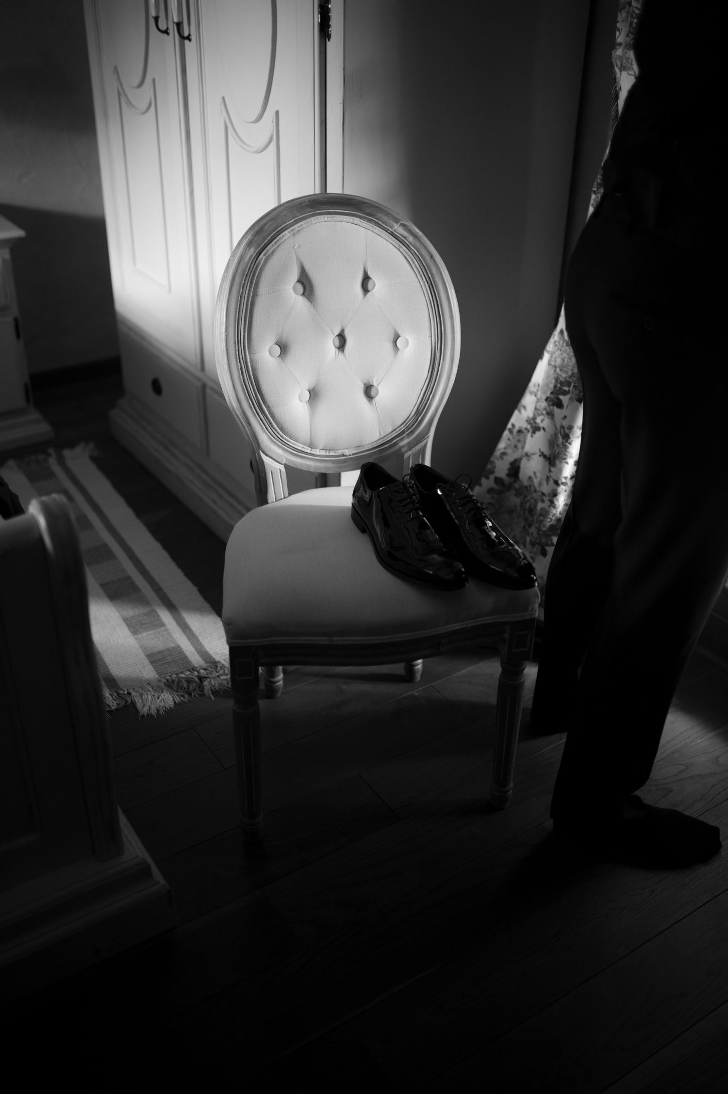 Black and white photo of elegant tufted chair with dress shoes, beside a wardrobe in a formal room.