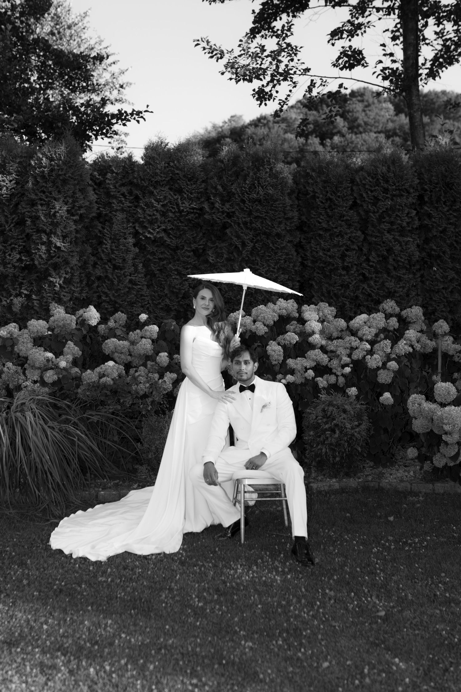 Black and white photo of elegant bride and groom posing in garden with hydrangeas, groom seated under parasol.