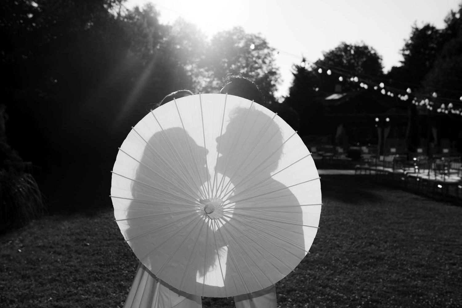 Black and white photo of couple kissing behind a white parasol umbrella at an outdoor wedding reception.