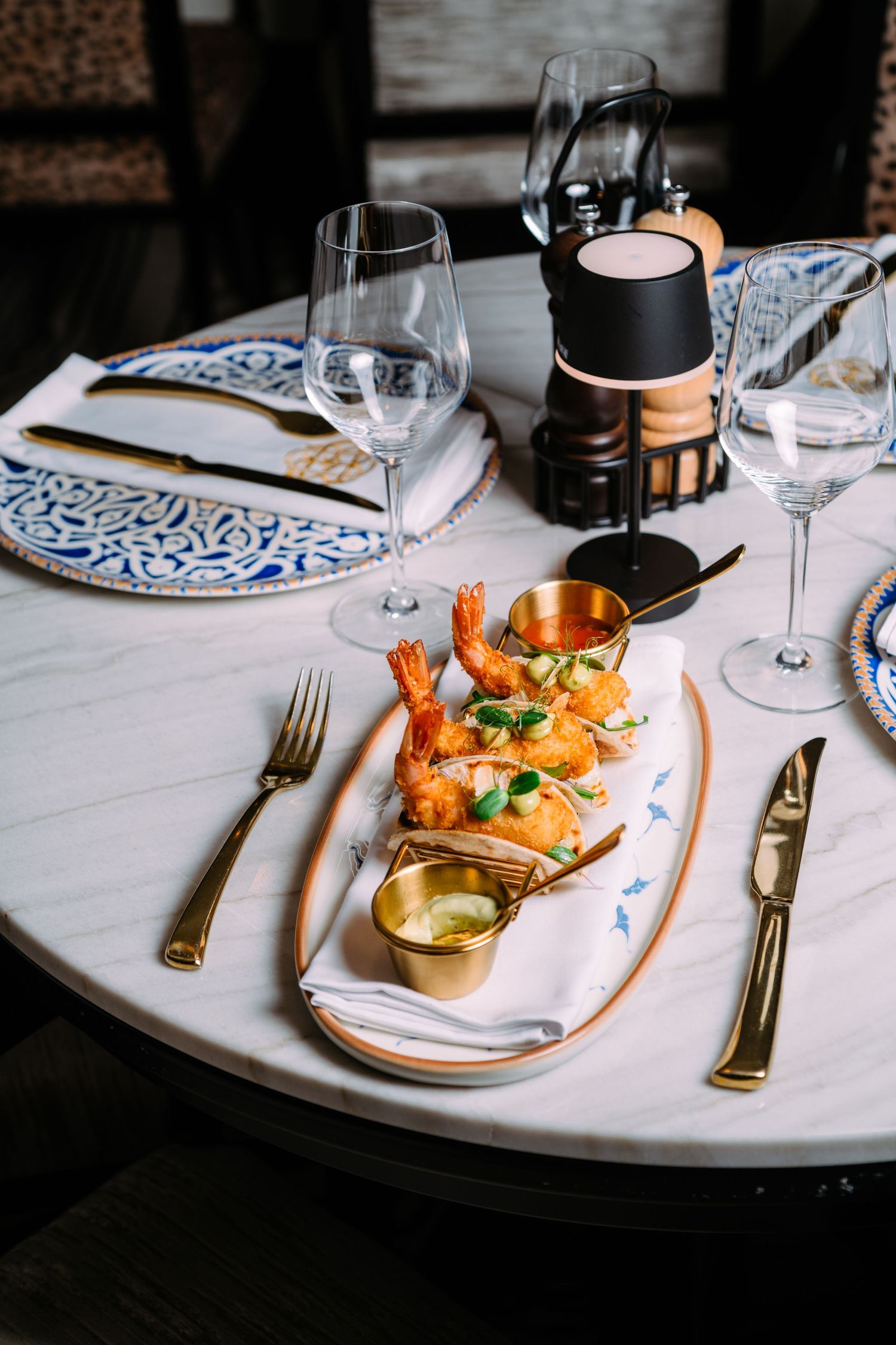 Elegant table setting with copper dish of prawns, decorative plates, wine glasses, and gold cutlery on white tablecloth.