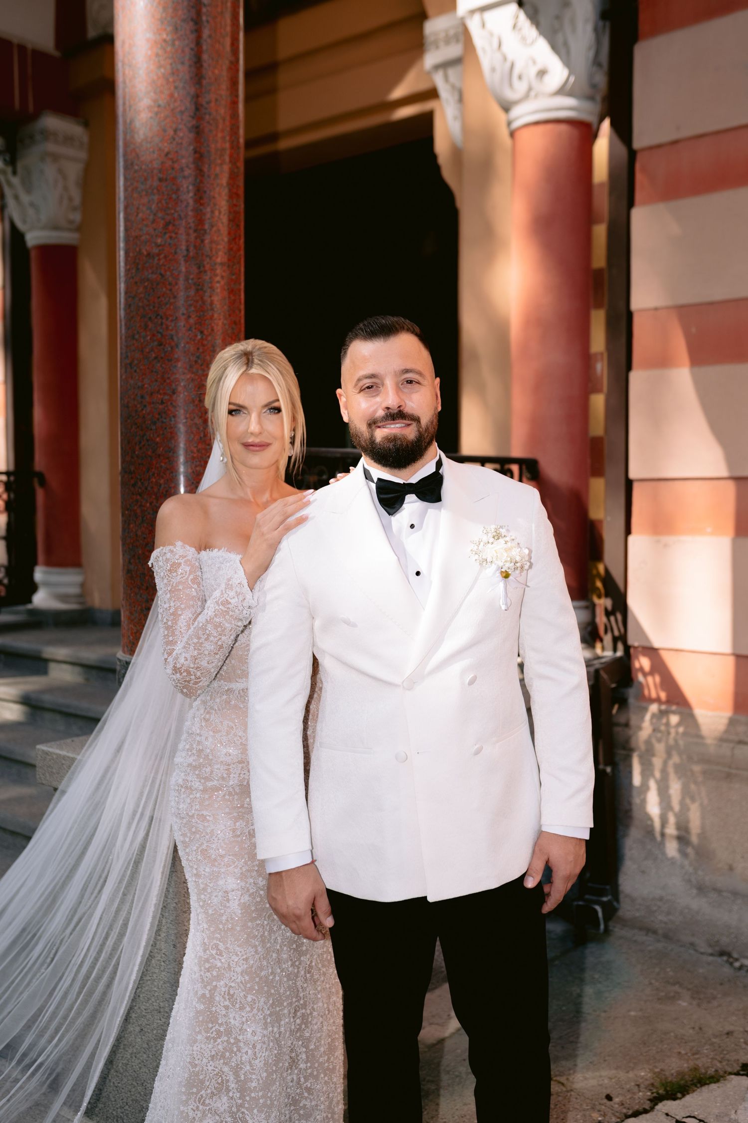 Wedding couple in formal attire posing against architectural columns.