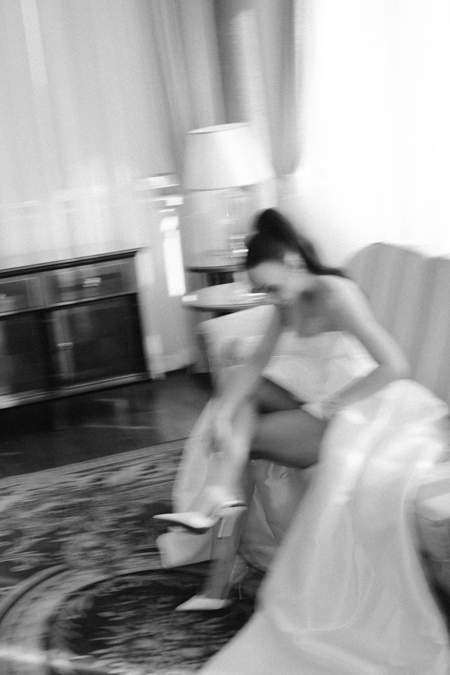 Black and white blurred photo of a bride in a white dress and heels sitting in an elegant room.