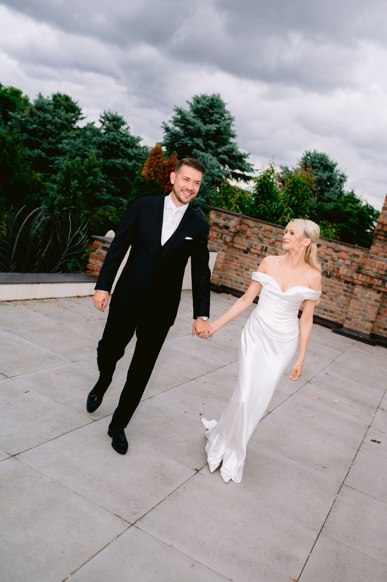 A smiling bride and groom hold hands walking outdoors, she in an off-shoulder white gown, he in a black tuxedo.