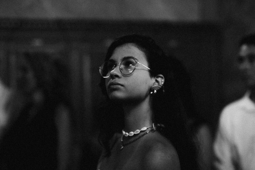 Black and white portrait of a young woman wearing round glasses and a necklace, looking upward thoughtfully.