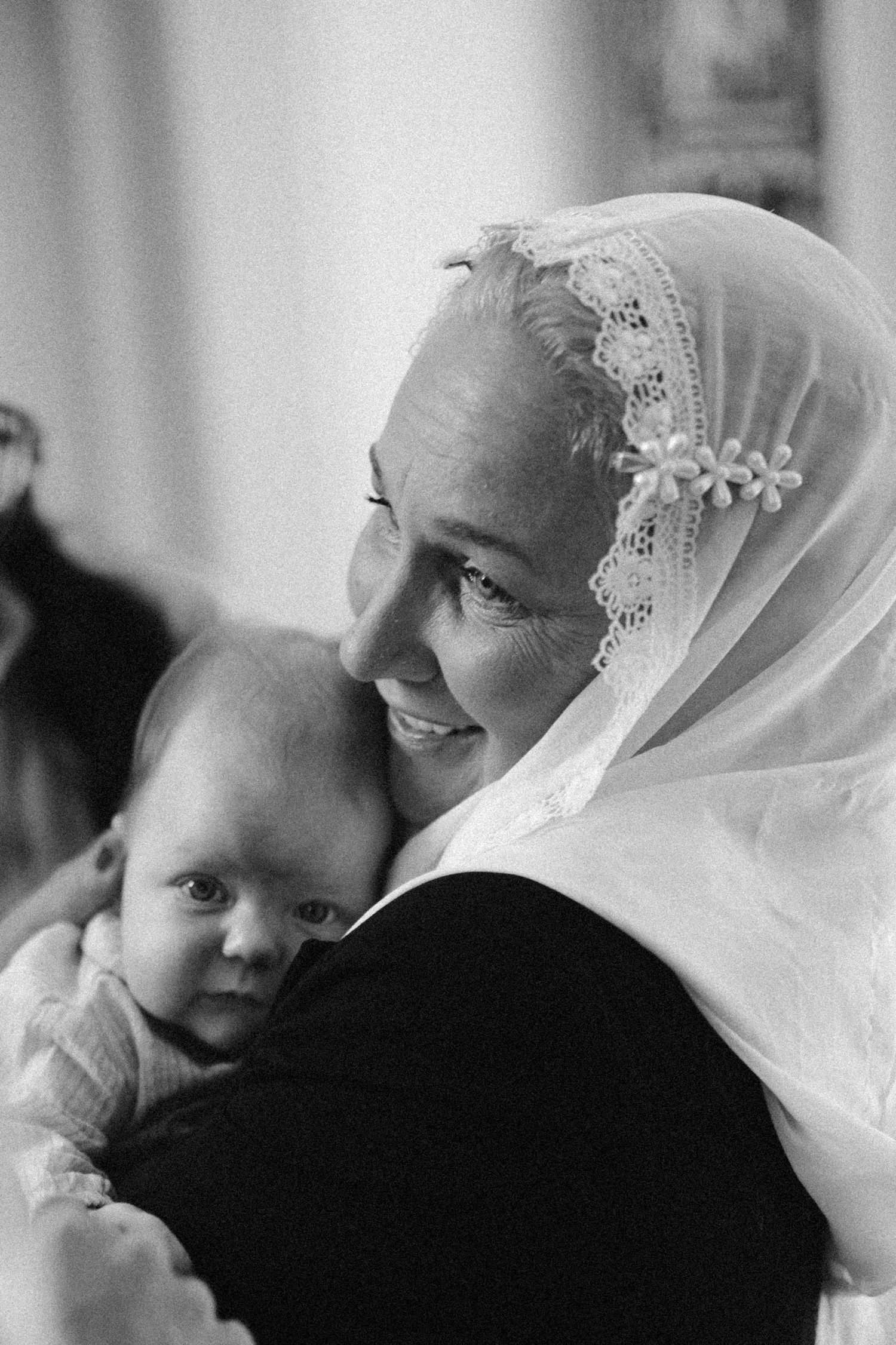 Black and white photo of a smiling elderly woman in a lace headscarf tenderly holding a newborn baby.