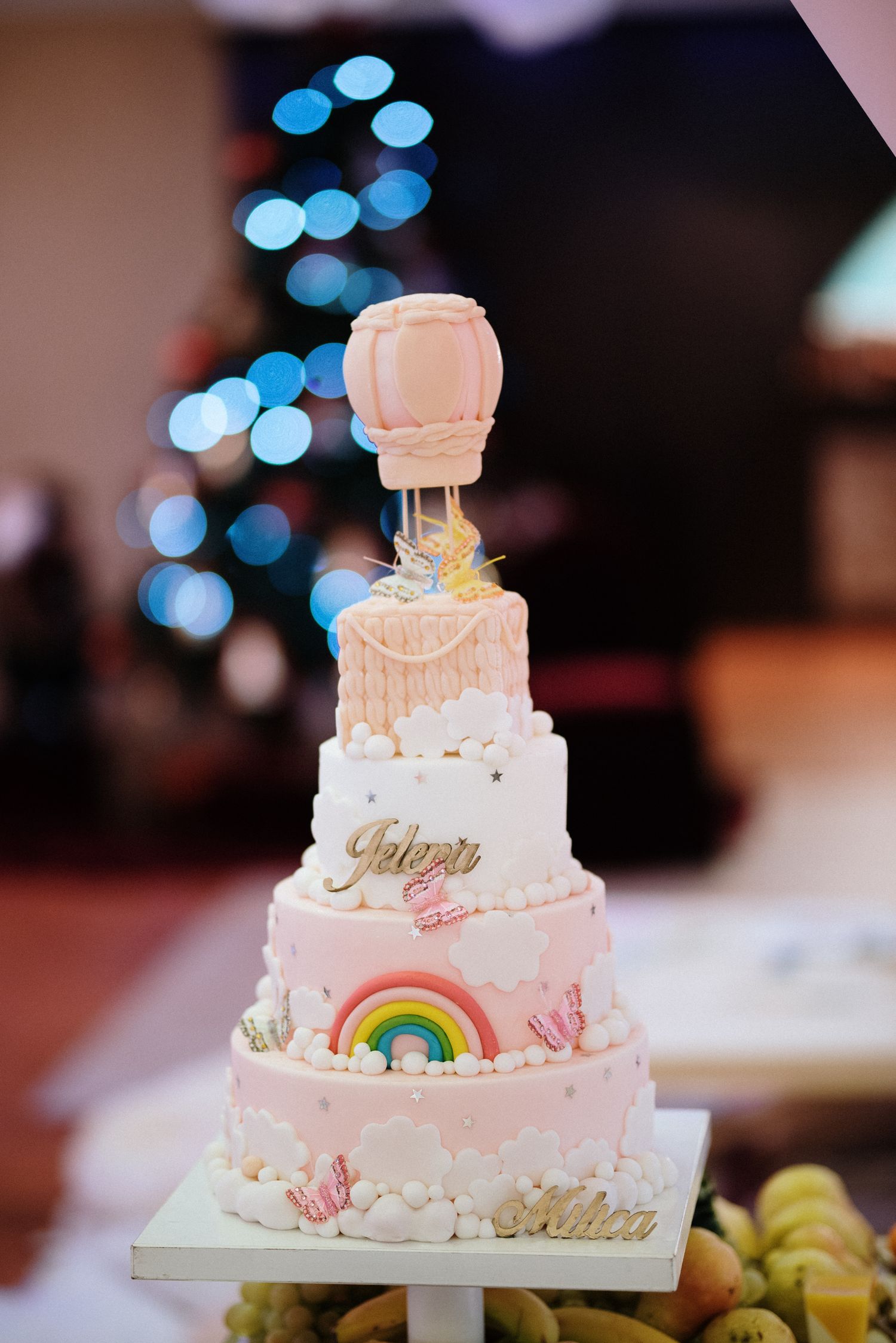 A elegant four-tier pink and white birthday cake with rainbow decor and hot air balloon topper.
