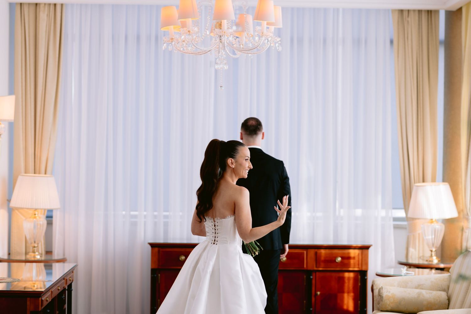 A wedding couple stands in an elegant room with white curtains and chandeliers during their first dance.