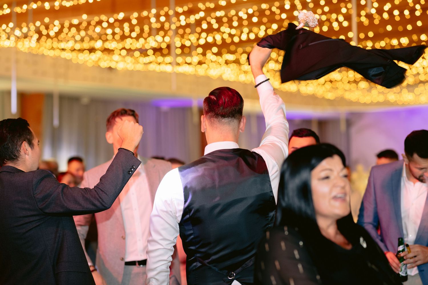 Wedding guests dance and celebrate on a dimly lit dance floor decorated with warm glowing string lights overhead.