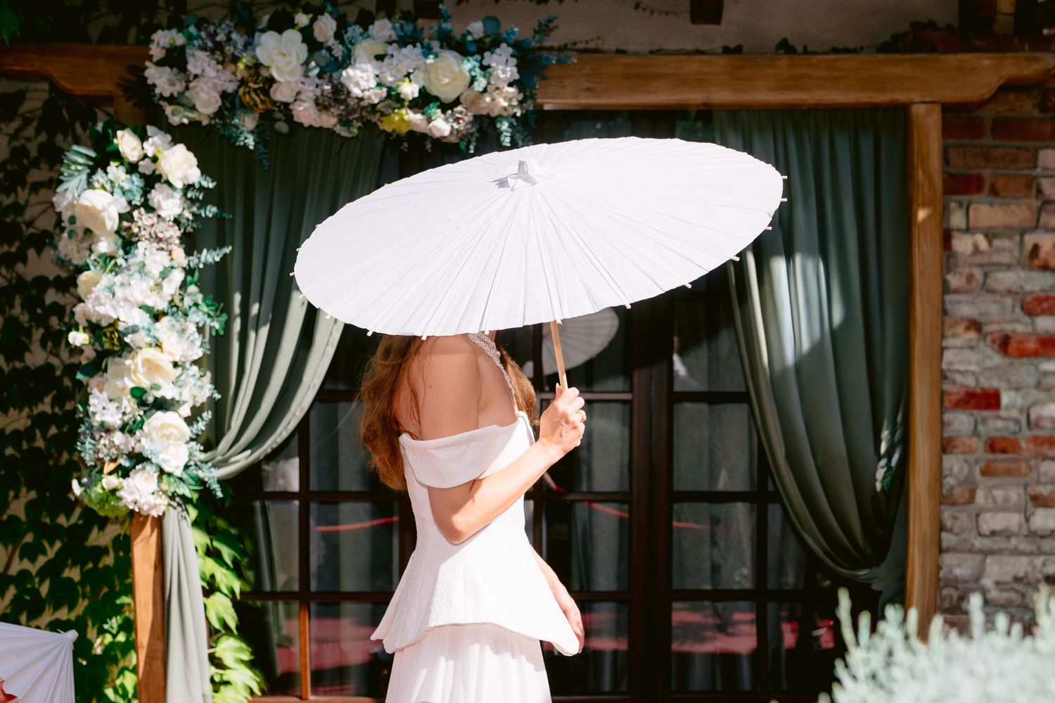 Bride in white dress holding elegant white parasol umbrella at floral wedding arch with green drapes.