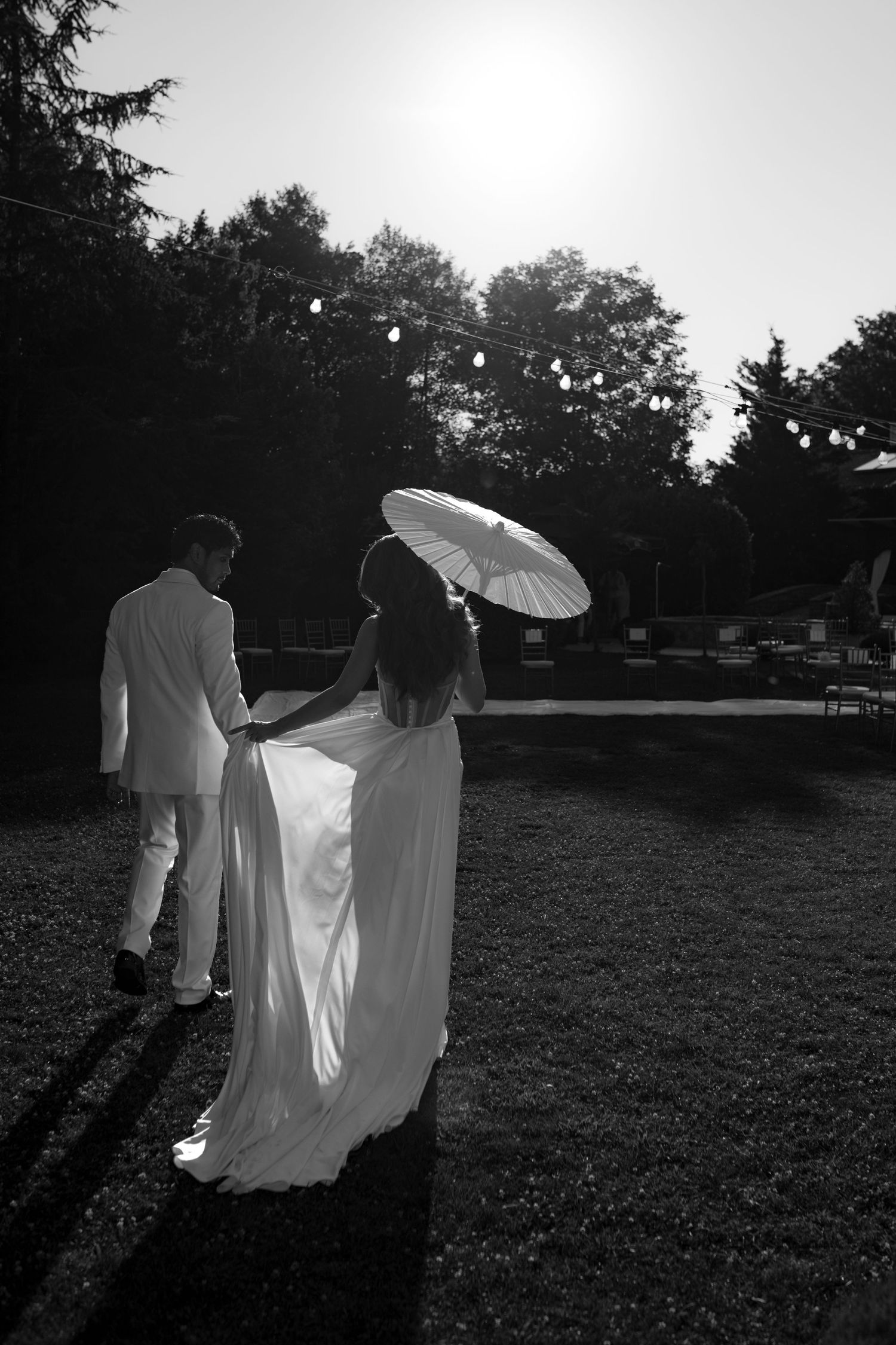 Black and white photo of bride and groom walking outdoors, bride holding parasol in flowing gown, backlit by sun.