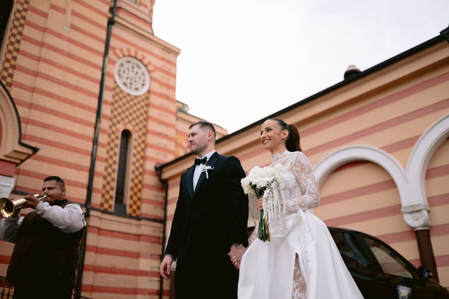 Wedding party celebrates joyfully outside a pink and peach striped building with arched architecture.
