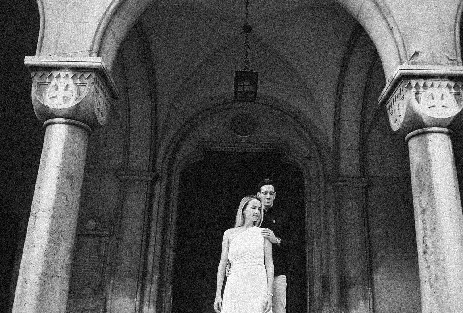 A couple in wedding attire poses under stone archways in a dramatic architectural setting.