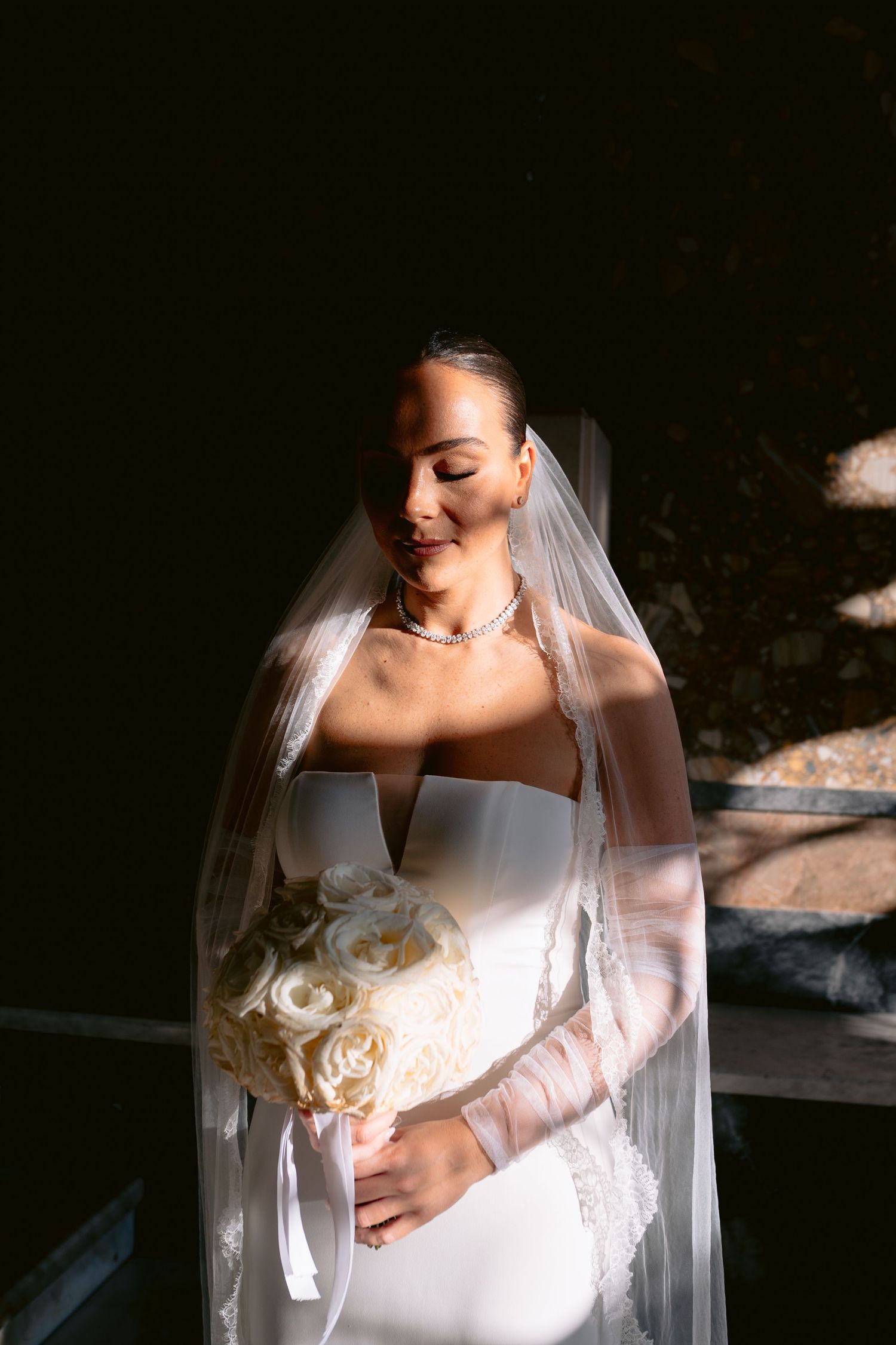 Elegant bride in a white strapless gown and long veil holds a bouquet of white roses in dramatic lighting.