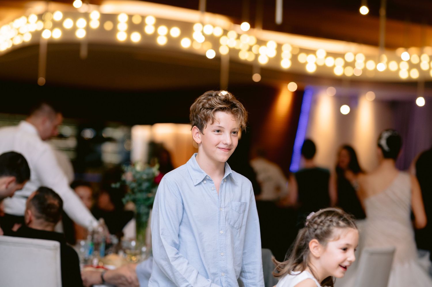 A smiling boy in a blue shirt at an elegant wedding reception with fairy lights and guests in the background.