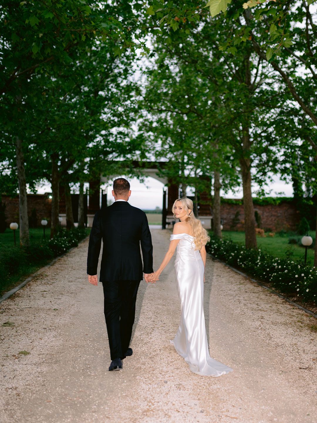 A bride and groom walk hand-in-hand down a tree-lined gravel path, the bride in a silver off-shoulder gown.