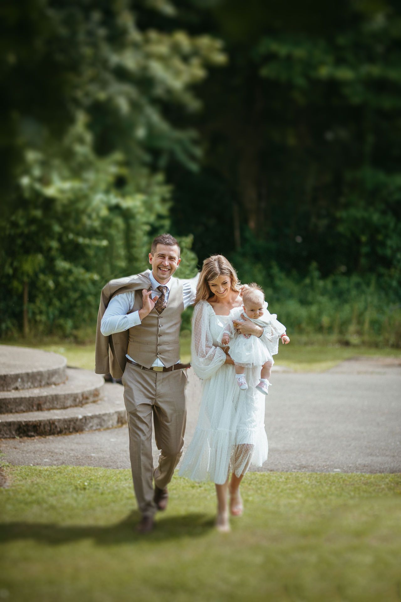 Happy couple in wedding attire walking outdoors, woman holding baby, man carrying jacket, lush green trees background.