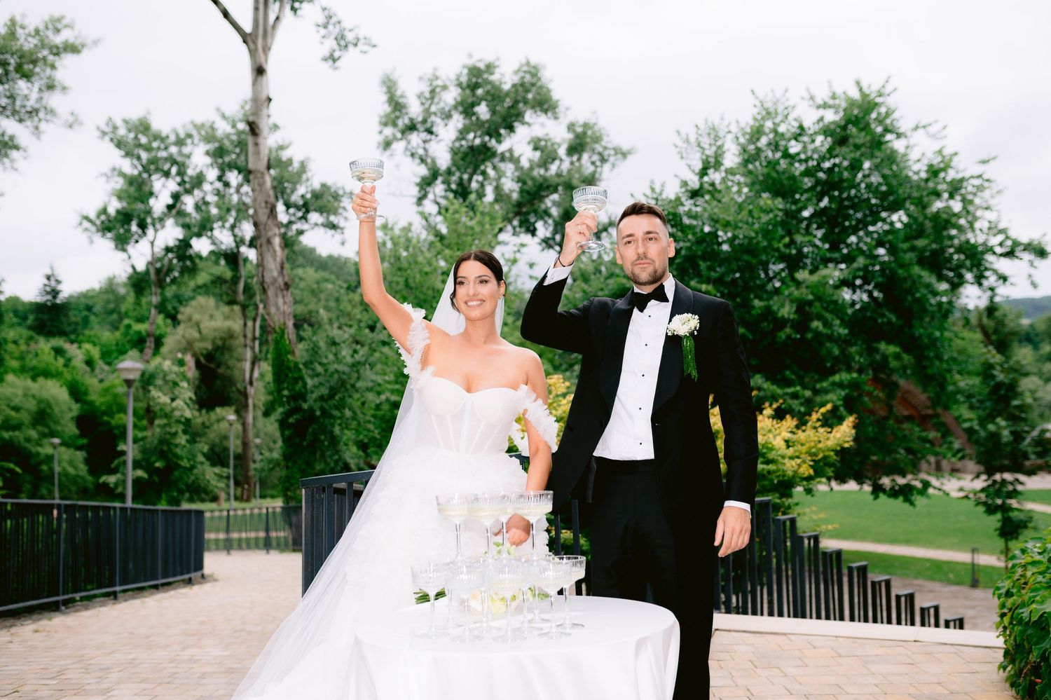 Bride and groom toasting with champagne glasses outdoors at elegant wedding venue surrounded by lush greenery.