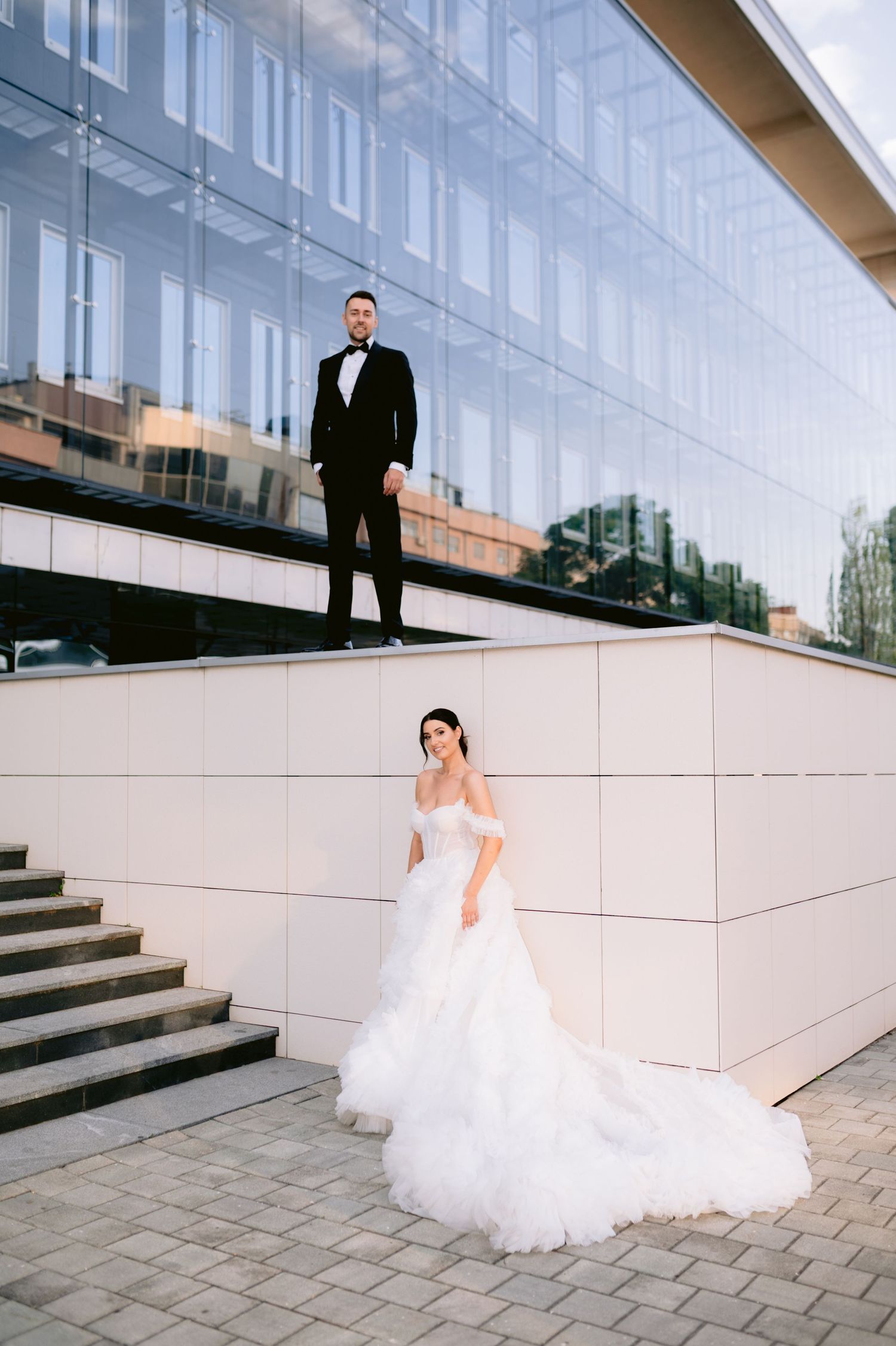 Elegant wedding couple posing outside modern glass building, bride in white off-shoulder gown, groom in black tuxedo on elevated platform.