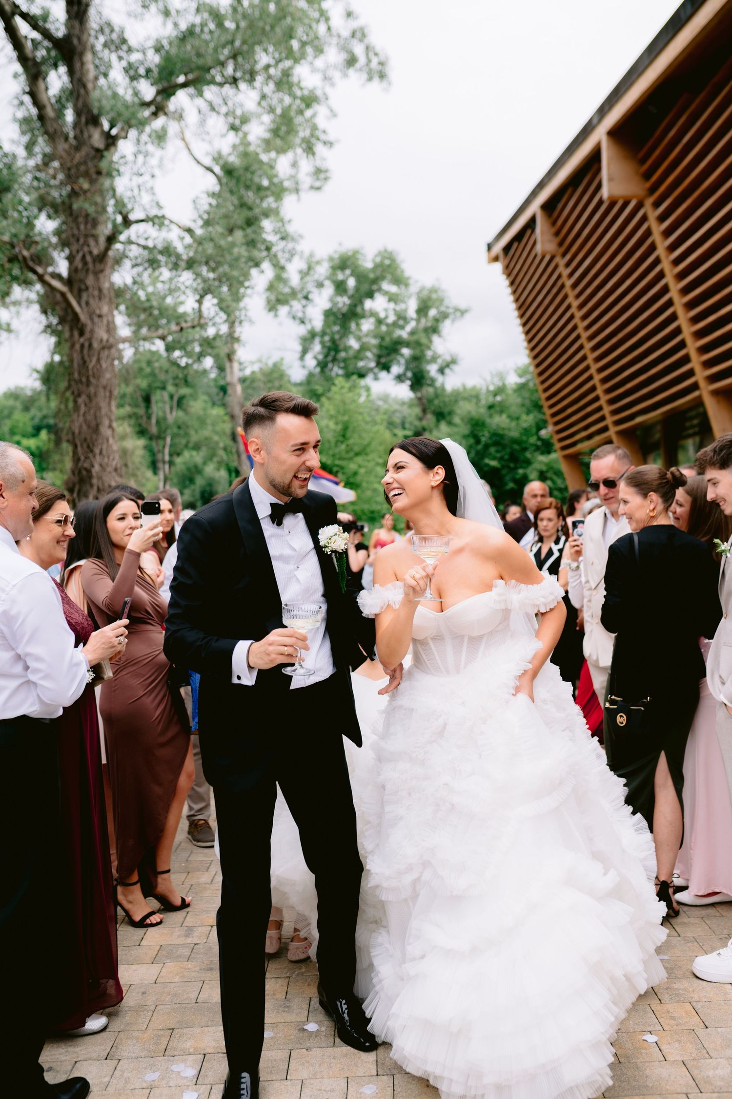 Joyful bride in ruffled off-shoulder gown and groom in tuxedo walk through cheering wedding guests outdoors.