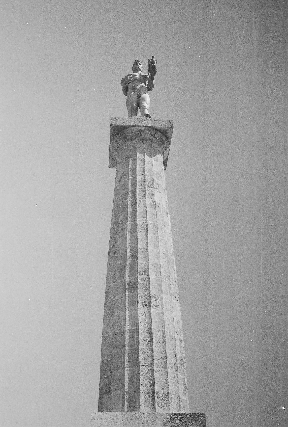 A tall stone column monument with a statue on top photographed from below against a plain sky.