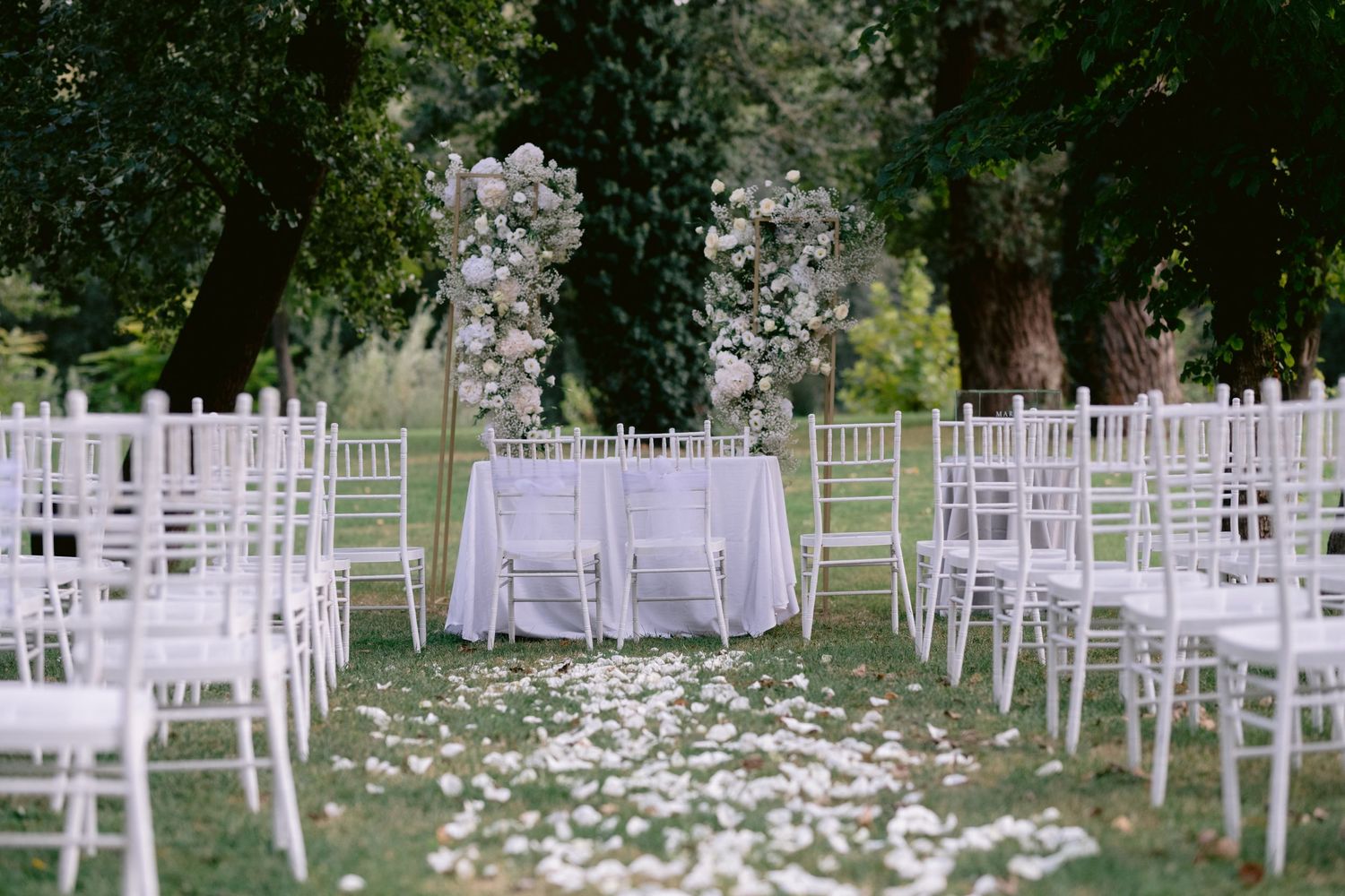 Elegant outdoor wedding ceremony setup with white chiavari chairs, rose petal aisle, and floral arch on lush green lawn.
