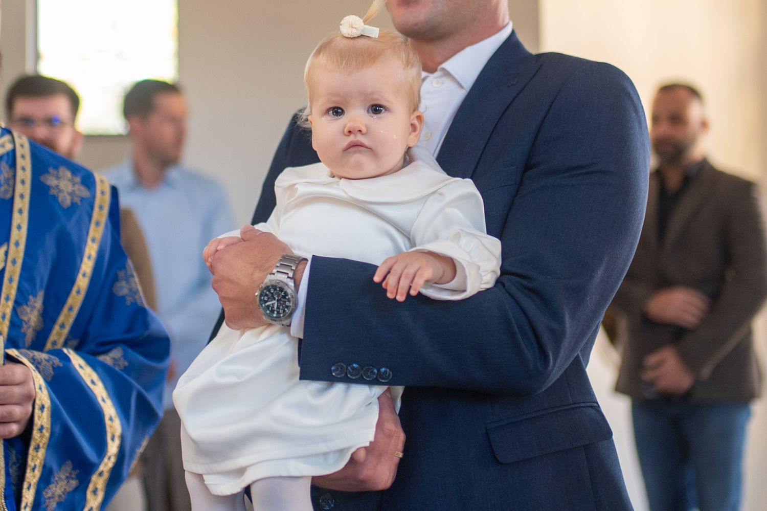 A baby in a white dress and bow is held by a man in a navy suit during a church ceremony, with guests standing in the background.