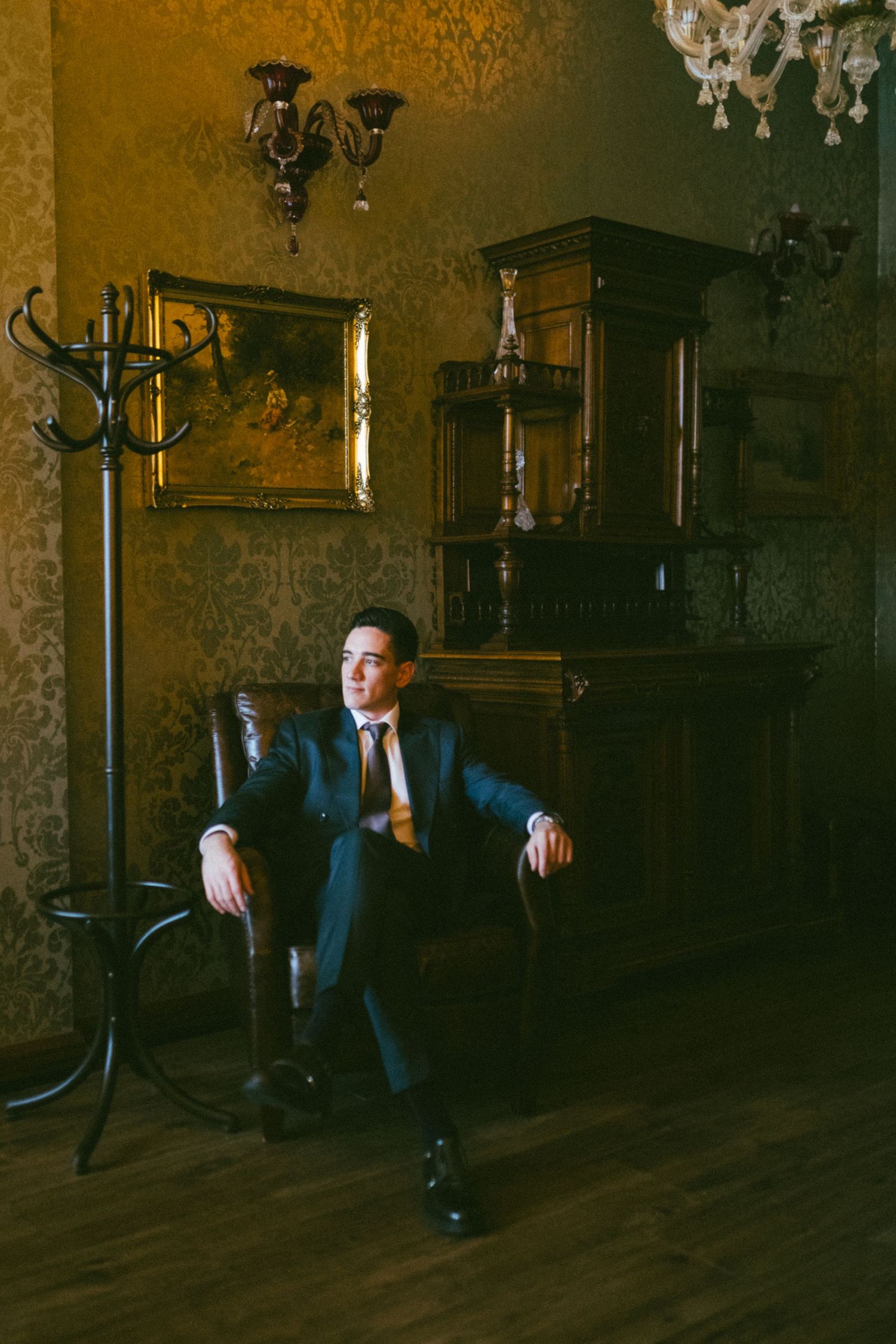 A person in a dark suit performs a breakdancing move on a hardwood floor beneath crystal chandeliers.