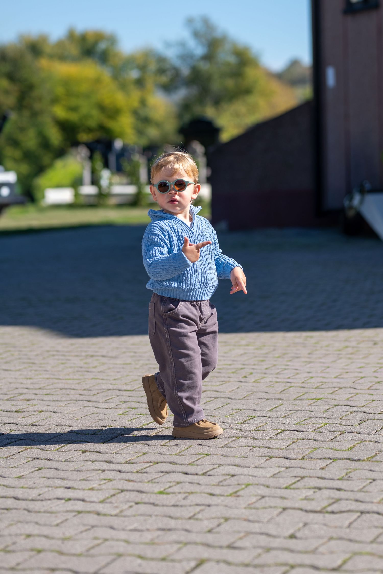 Toddler in round sunglasses, blue knit sweater and gray pants walks on a sunlit cobblestone path, gesturing with one hand.