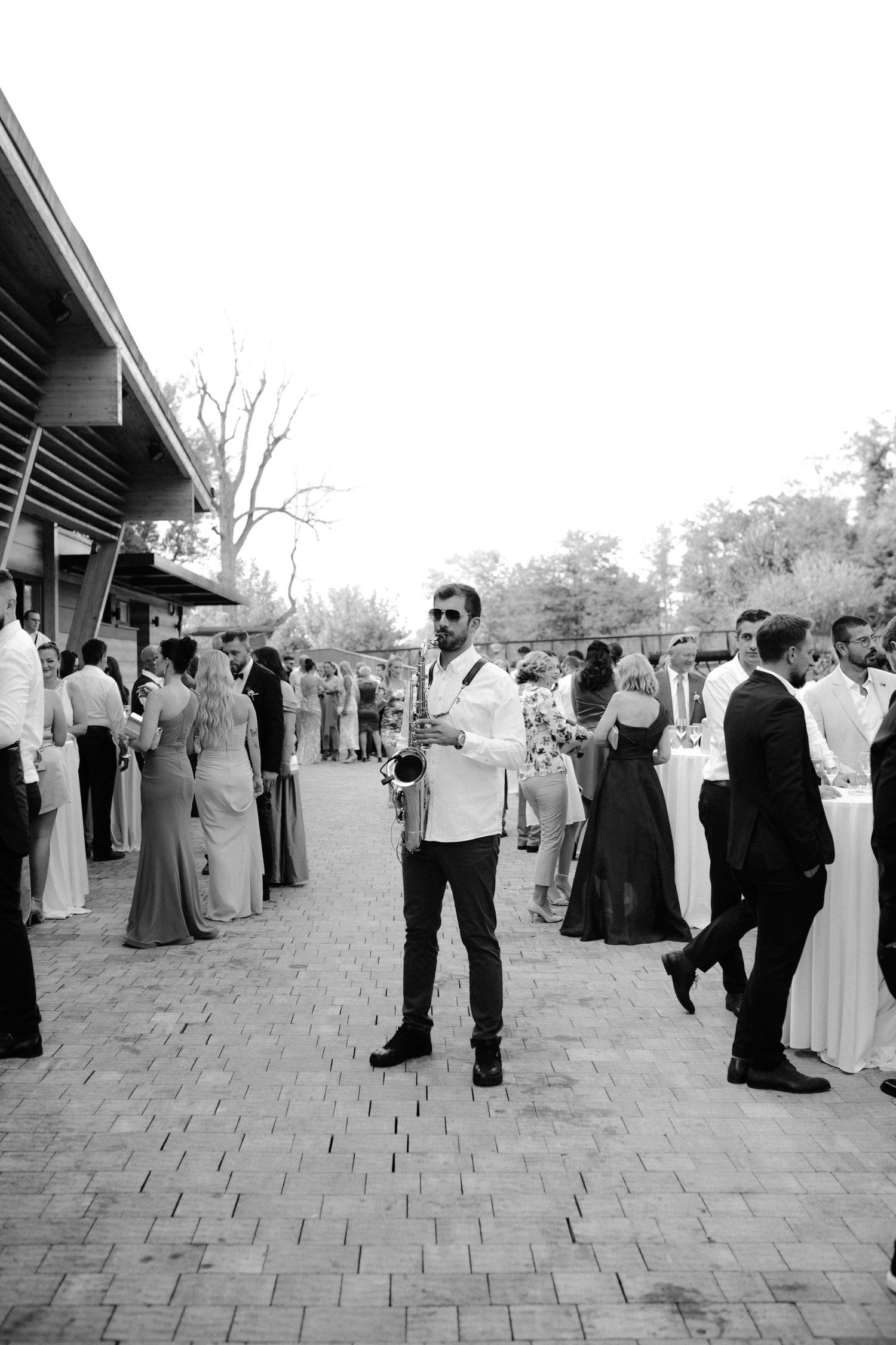Black and white photo of a man in a white jacket standing at an outdoor wedding reception with guests mingling.