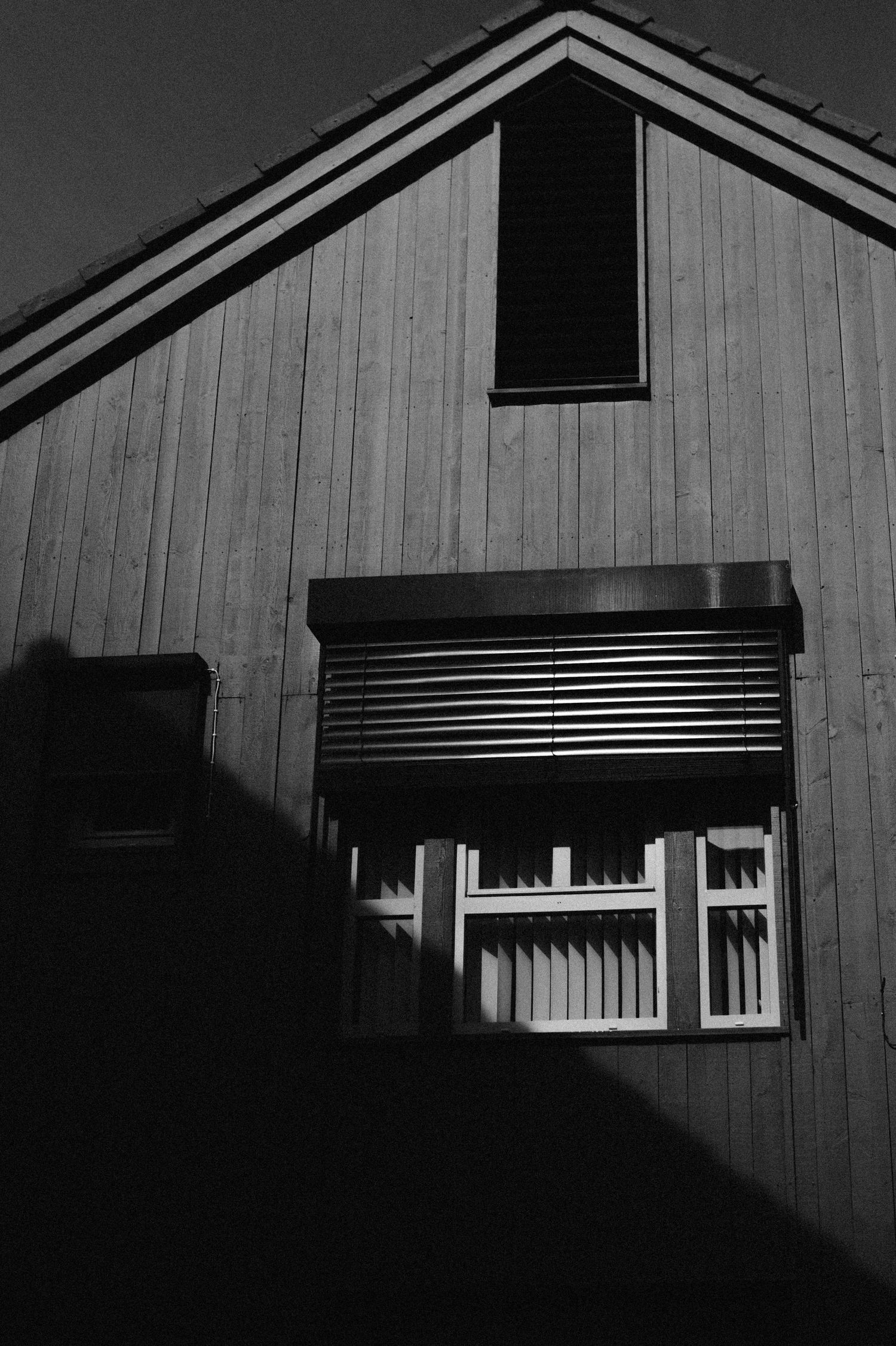 Black and white architectural photo of a wooden house exterior with a window and rolling shutter casting dramatic shadows.