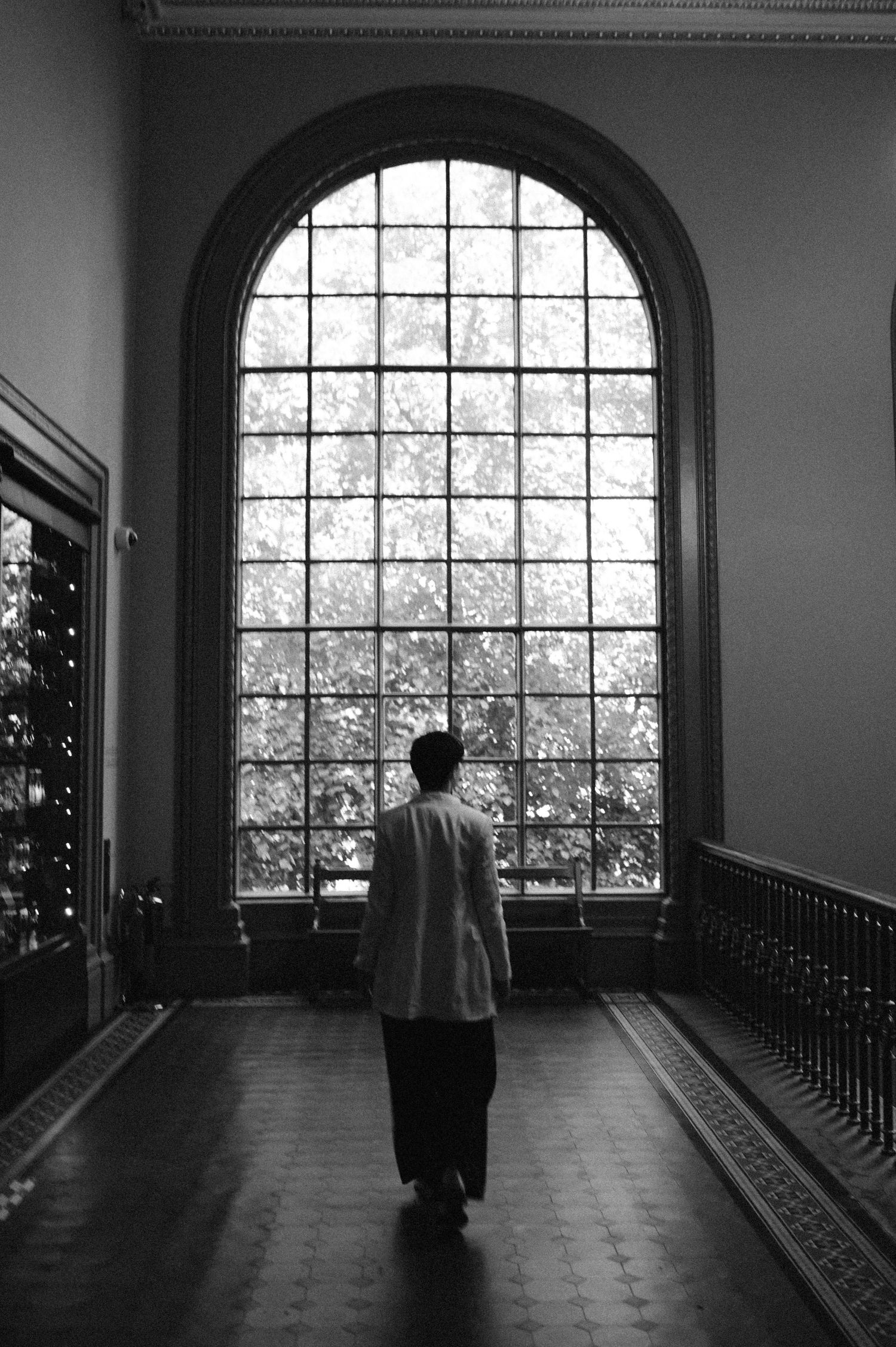 A silhouetted figure stands in a dark hallway with large arched windows creating a dramatic black and white composition.