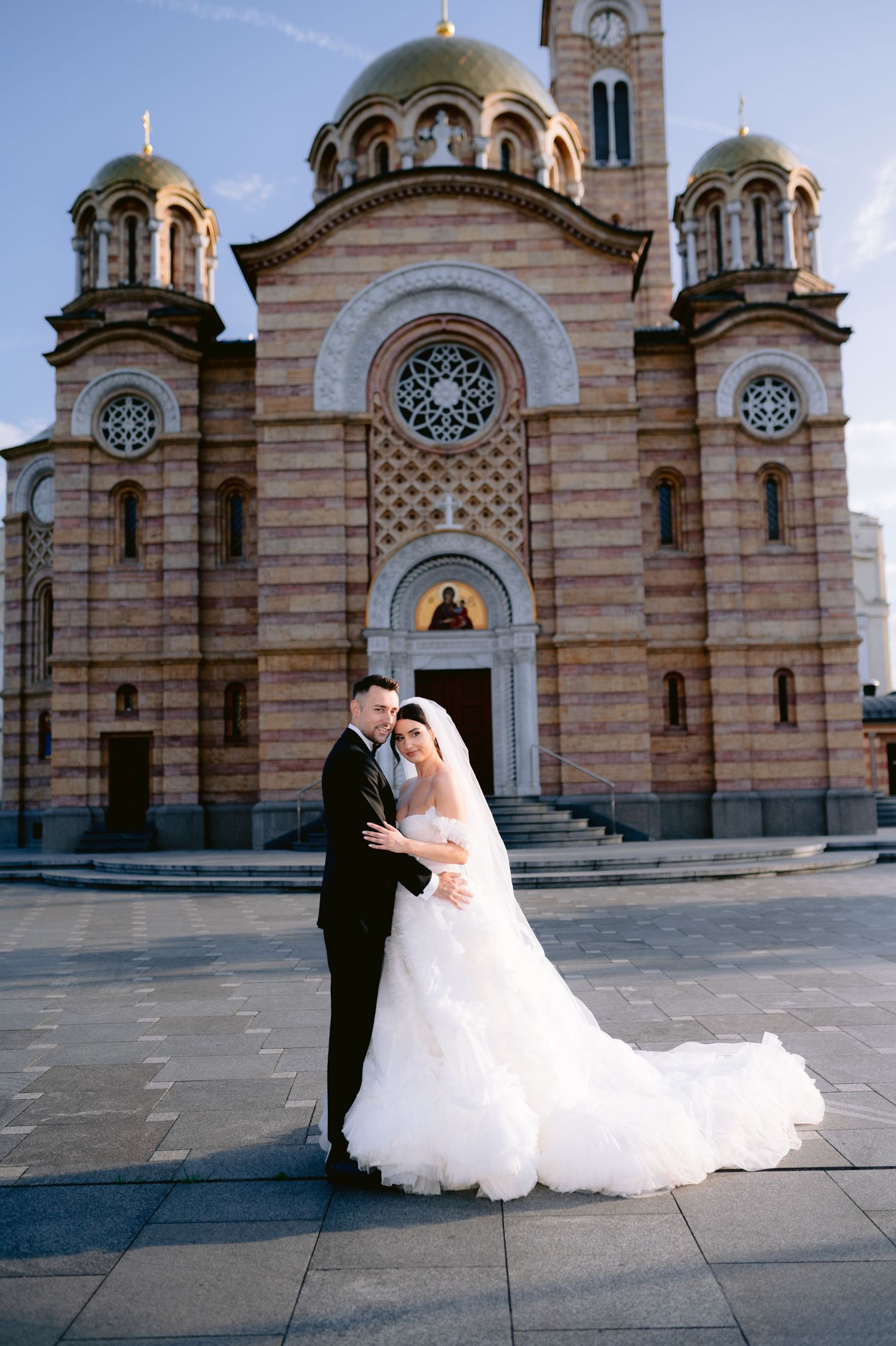Bride and groom embracing in front of a grand Orthodox church with golden domes on their wedding day.