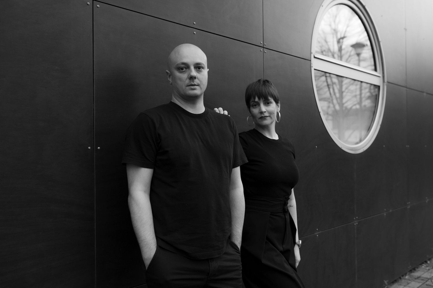 A black and white photograph shows two people in dark clothing standing against a wall with a round window.