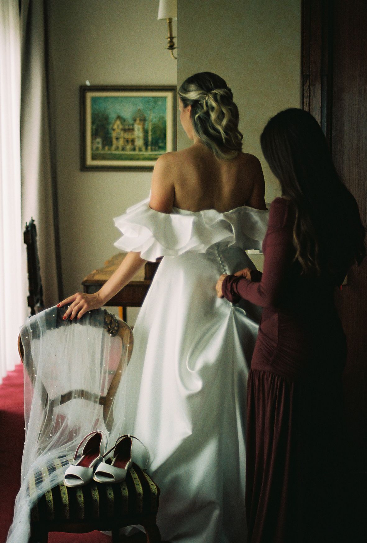 A bride in a white off-shoulder gown gets help with final preparations while standing in a dimly lit room.