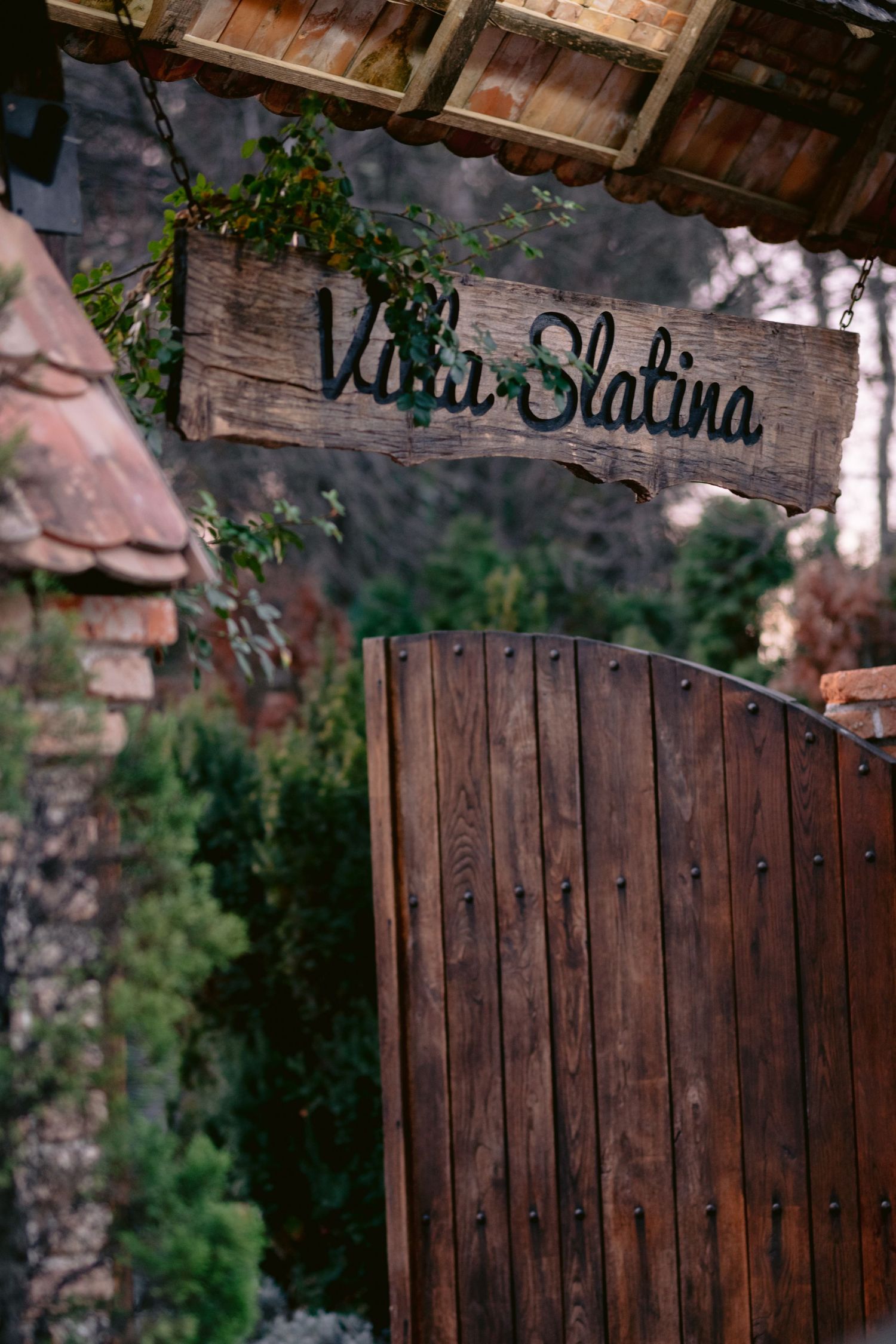 Rustic wooden gate entrance with 'Villa Slatina' sign mounted above, surrounded by stone walls and green foliage.