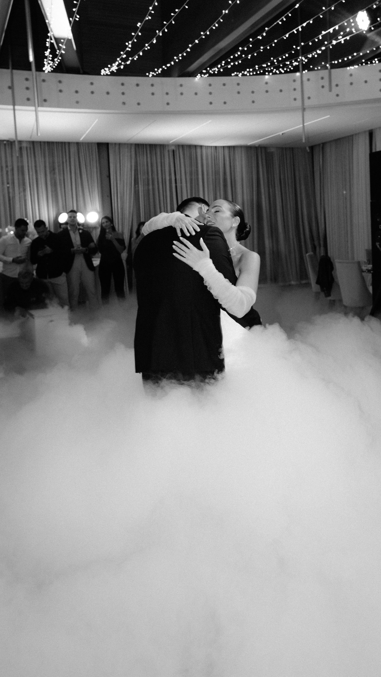 Bride and groom share romantic first dance surrounded by dramatic fog machine clouds at elegant wedding reception.