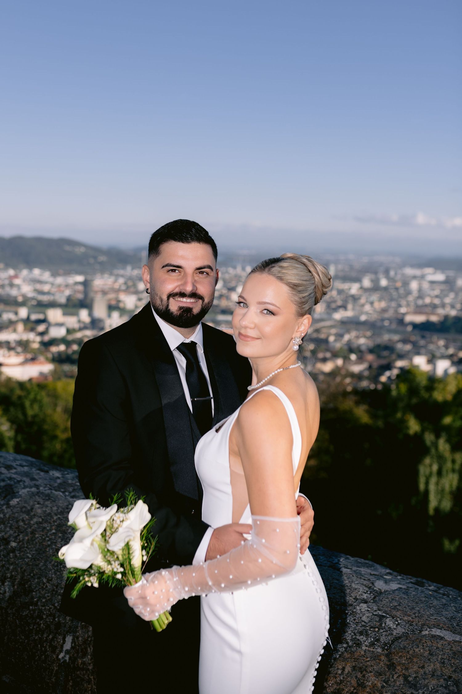 Wedding couple poses against cityscape backdrop on hilltop overlook.