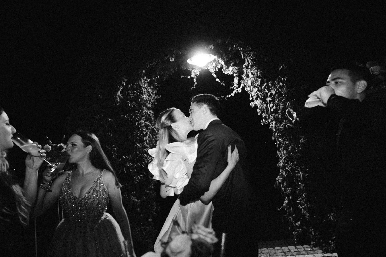 Two people share a romantic kiss at a nighttime wedding celebration with sparkling lights in the background.