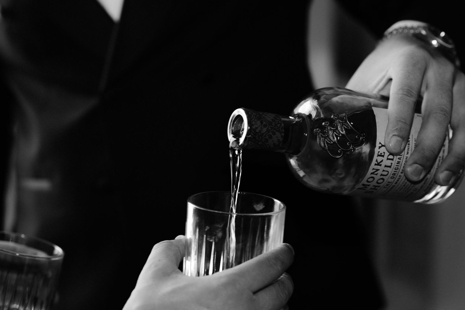 Black and white photo of someone pouring a drink into a glass with a ring visible on their hand.