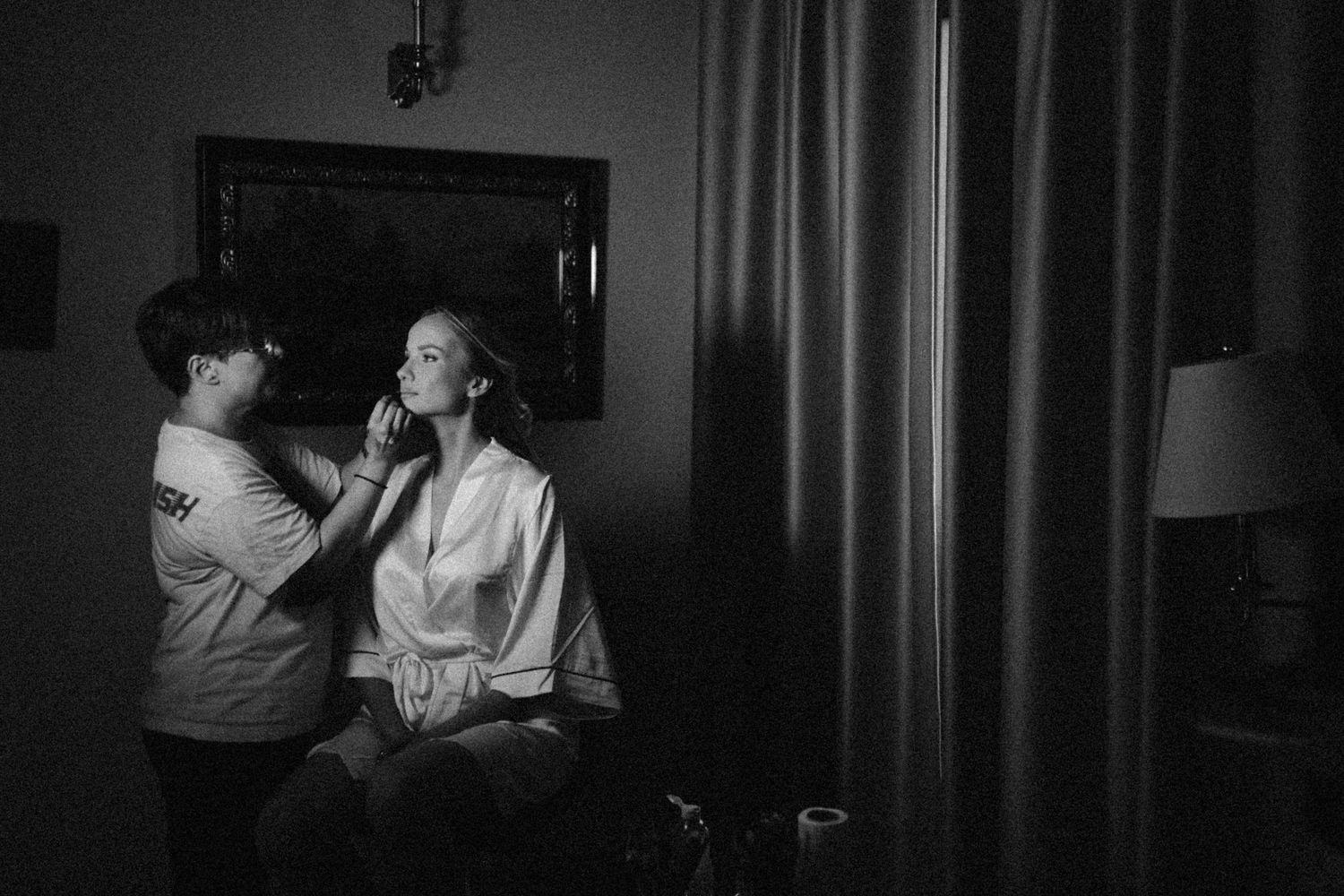 A black and white photo shows makeup being applied in a dimly lit room with a curtain and lamp visible.